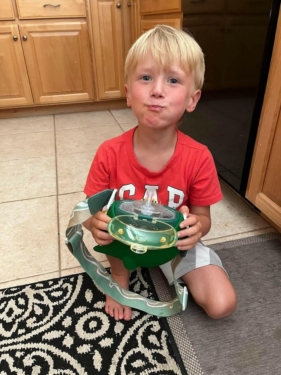 Young boy in a red shirt holding a dino-rific-dinosaur-kids-water-bottle-with-straw in a kitchen.